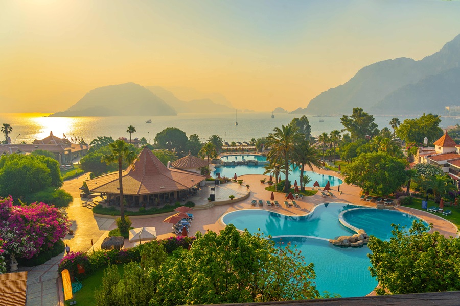 Aerial view of a tropical resort with winding blue pools, palm trees, and a sunlit bay at sunset.