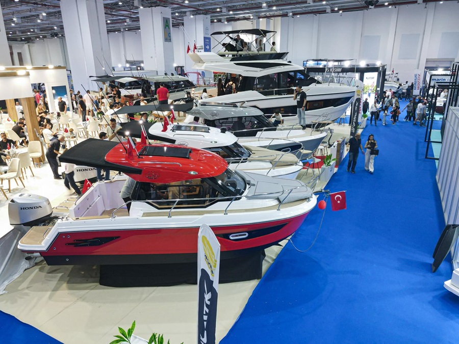 Indoor boat show featuring a red and black motorboat in the foreground, with white yachts on display and attendees walking the blue-carpet aisle.