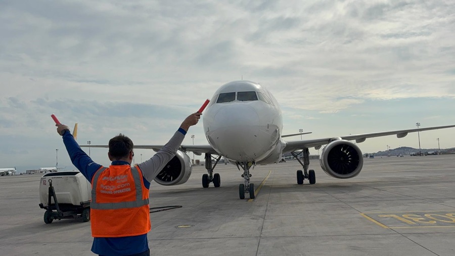 Ground crew member in orange safety vest signals an arriving airliner with red marshalling wands on a tarmac.
