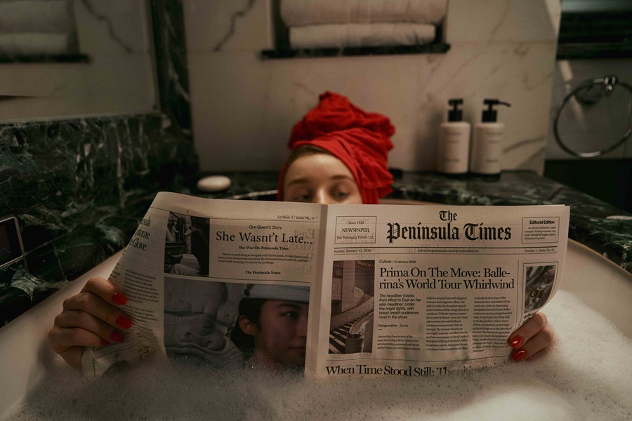 Person with a red towel on their head reading a newspaper in a bubbly bathtub.