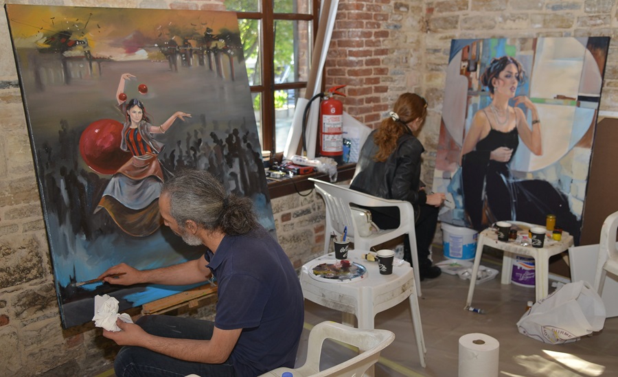 Two artists in a brick-walled studio painting large canvases; foreground artist portrays a dancer with a red ball, background artist fills a circular portrait.