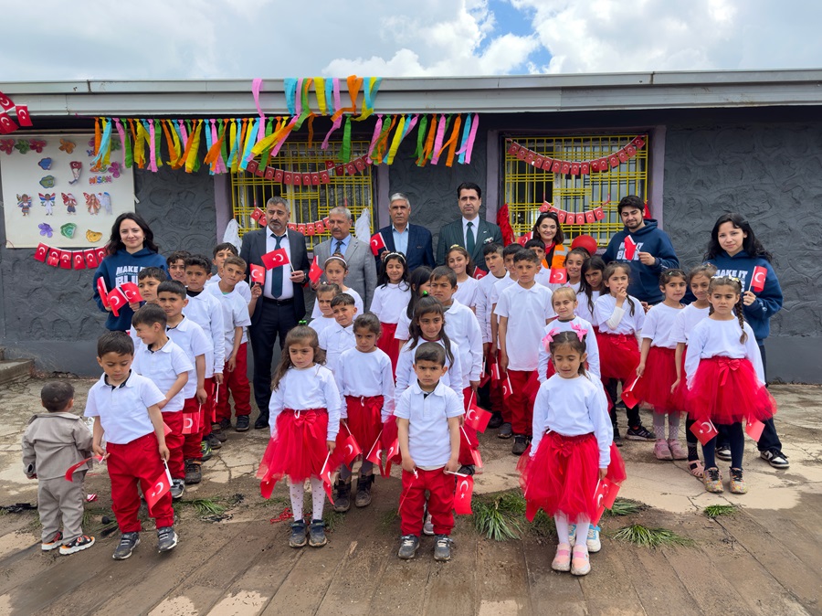 Group of children in white tops and red skirts, holding Turkish flags, posing with adults in front of a decorated school building.