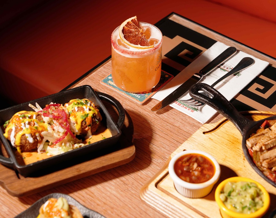 Orange cocktail with a citrus garnish on a coaster, next to a skillet of loaded tacos and dipping sauces on a wooden table.