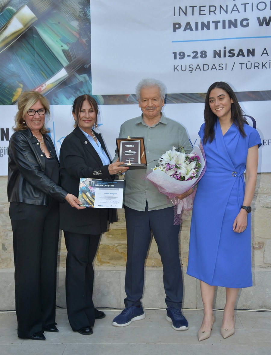 Group of four people posing at an award ceremony; elderly man center holds a plaque and bouquet as two women stand beside him, and a woman in a blue dress stands to the right, with a large banner in the background.