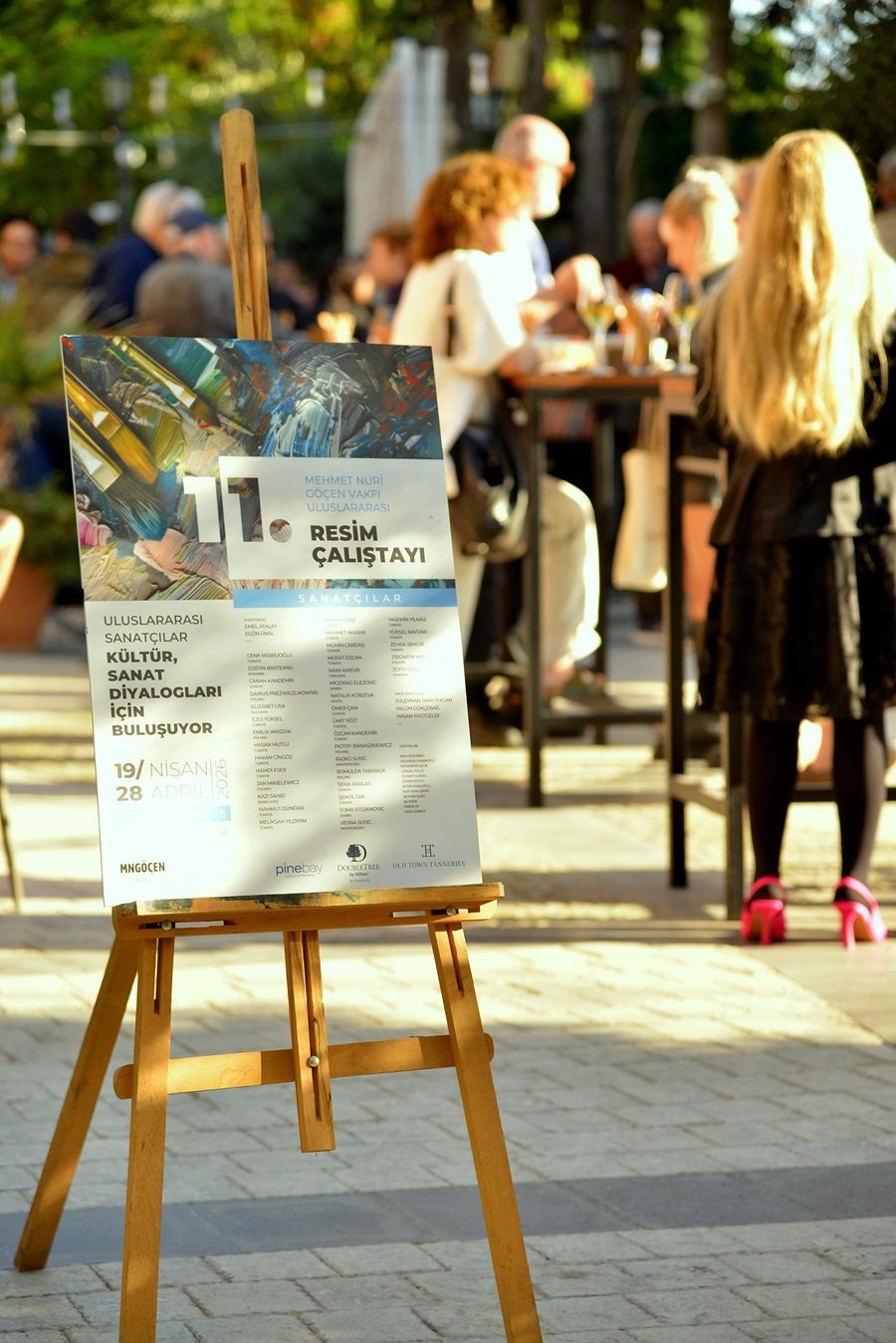 Poster on a wooden easel advertising an international painting workshop called 'Resim Çalıştayı' at an outdoor gathering with people chatting in the background.