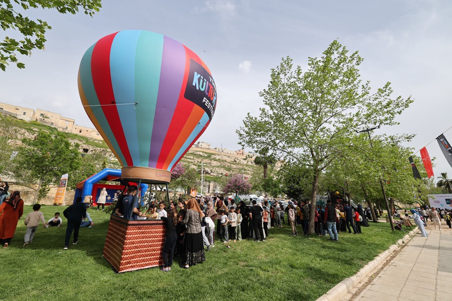 Colorful striped hot air balloon tethered in a grassy park, with people lining up near the basket and trees in the background.