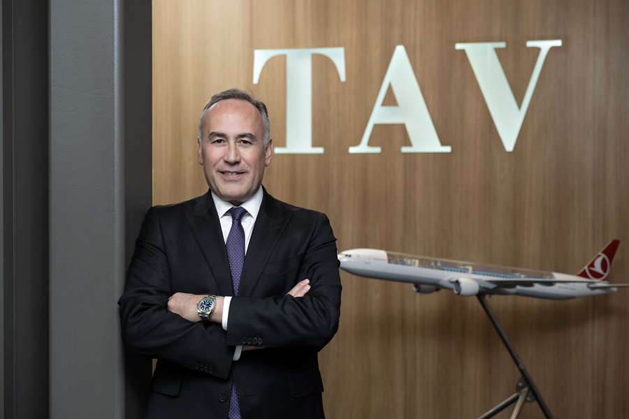 Businessman in a dark suit stands with arms crossed beside a small airplane model, in front of a wooden wall with 'TAV' lettering.