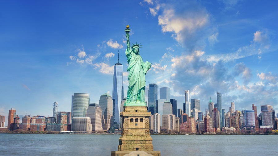Statue of Liberty on its pedestal with the Manhattan skyline and blue sky in the background, viewed from Liberty Island in the harbor.