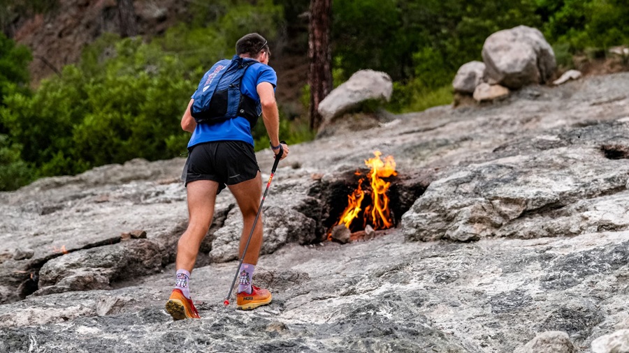 Hiker in a blue shirt with a backpack approaches a small campfire on rocky terrain.