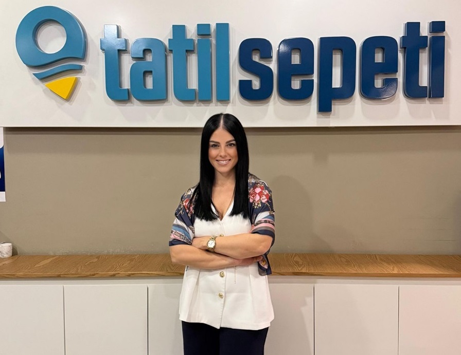 Smiling woman with long dark hair and a white top with patterned sleeves, arms crossed, standing in front of a sign that reads 'tatilsepeti'.