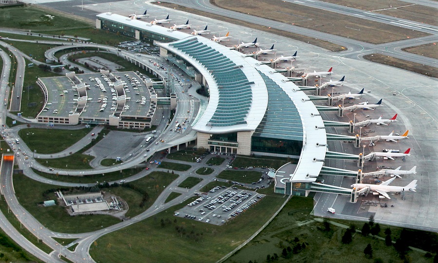Aerial view of a curved airport terminal with jet bridges attached to many airplanes parked at gates, surrounded by roads and a parking lot.