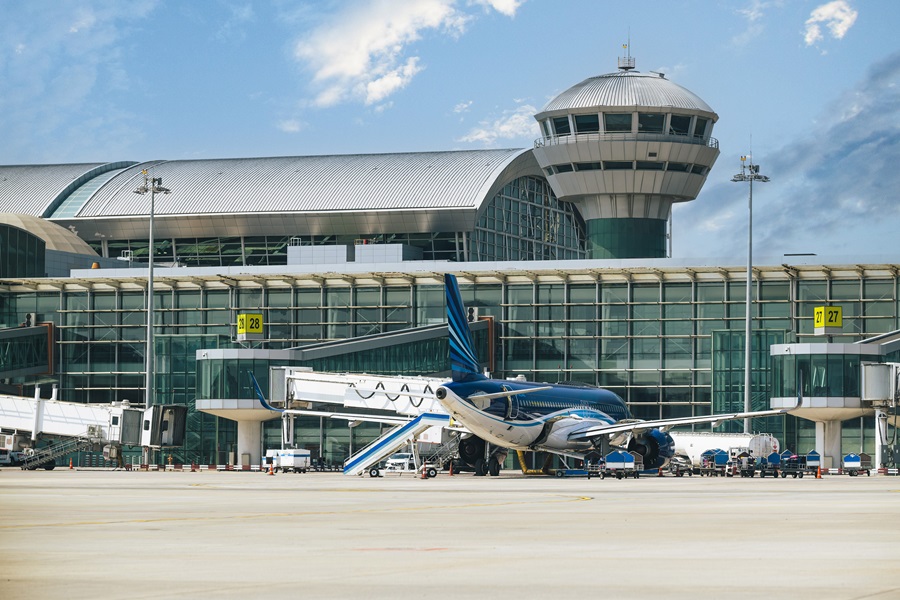Blue and white jet at a gate in front of a modern airport terminal with a circular control tower in the background on a sunny day.
