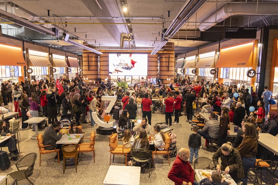 Crowded indoor dining area in a shopping center, with dozens of people eating at orange-upholstered tables under warm lighting.