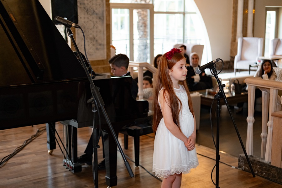 Young girl in a white dress sings into a microphone beside a grand piano, with an audience watching in a cozy recital room.