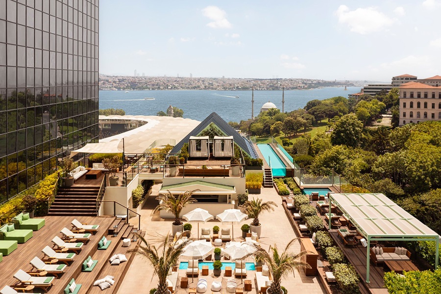 Rooftop pool and lounge area with palm trees overlooking a bay, a glass-walled building on the left, and distant cityscape.