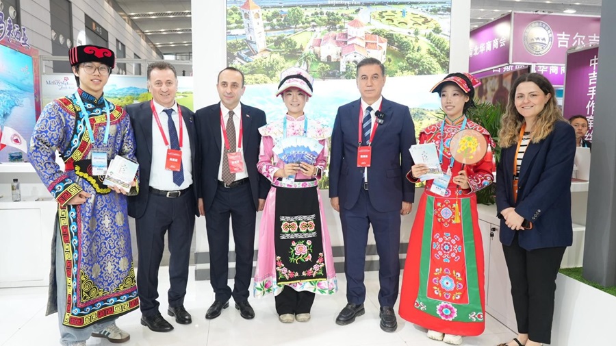 Group of seven people posing at a trade show; three wearing colorful traditional outfits, four in suits, holding brochures. A large scenic backdrop is behind them.