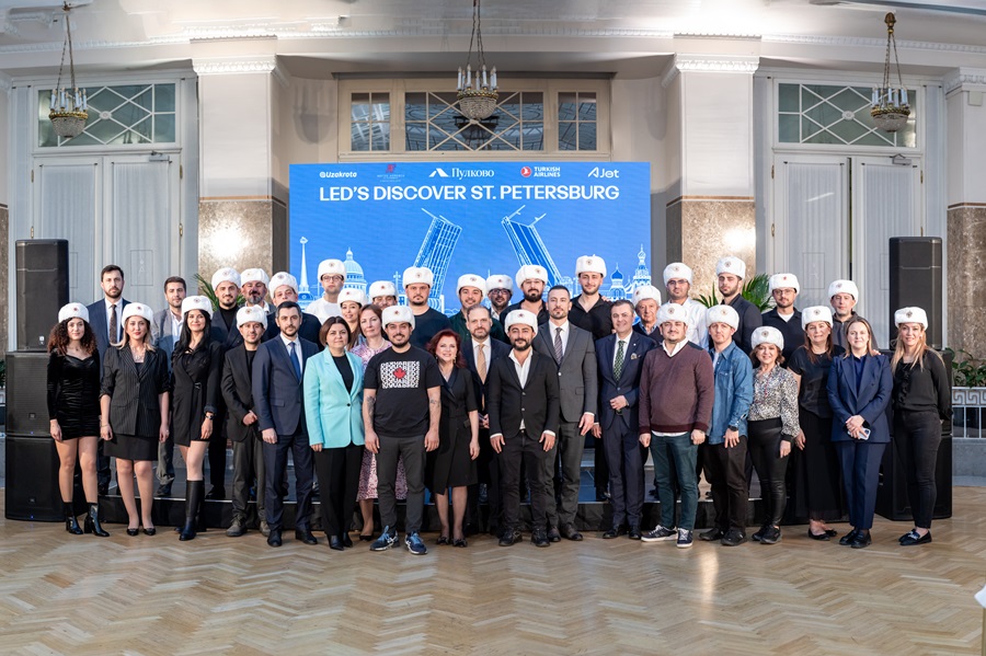 Group portrait of men and women in formal attire wearing white hats, posing at the LED's Discover St. Petersburg event in a grand hall with a blue banner behind them.