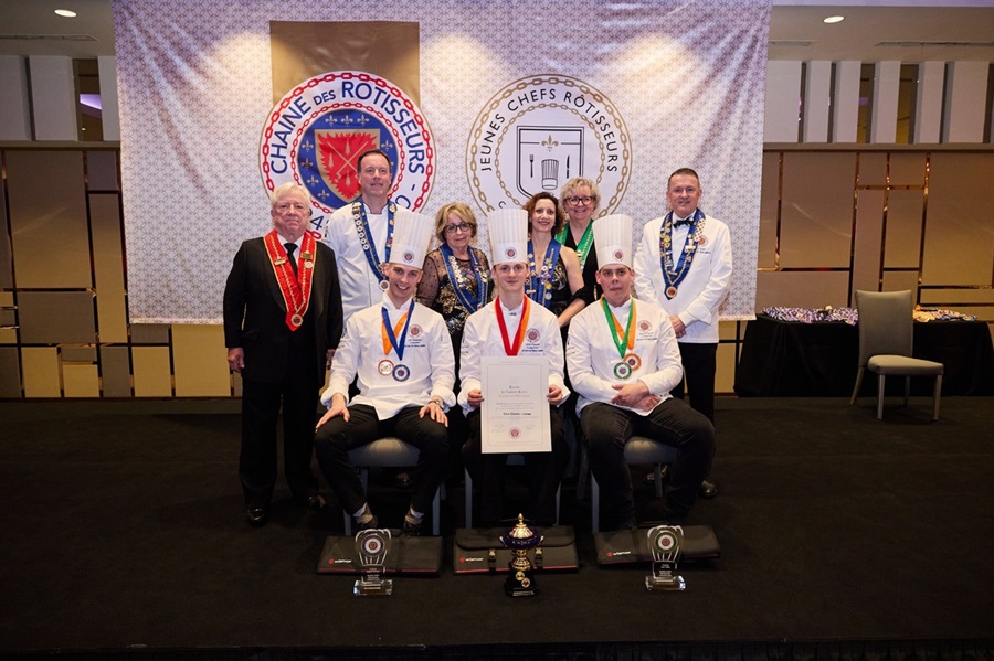Team of chefs on a stage posing with medals, certificates, and trophies at a formal culinary event.