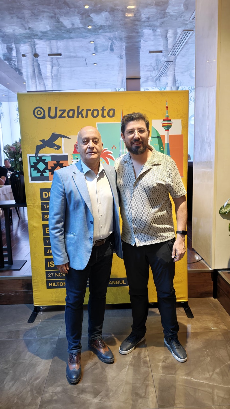 Two men pose for a photo in front of a yellow Uzakrota event banner in a modern venue with a colorful skyline graphic behind them.