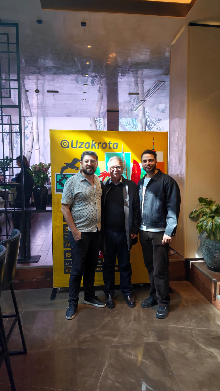 Three men posing indoors in front of a yellow Uzakrota banner; left man in a patterned shirt, center older man in a dark jacket, right man in a black leather jacket.