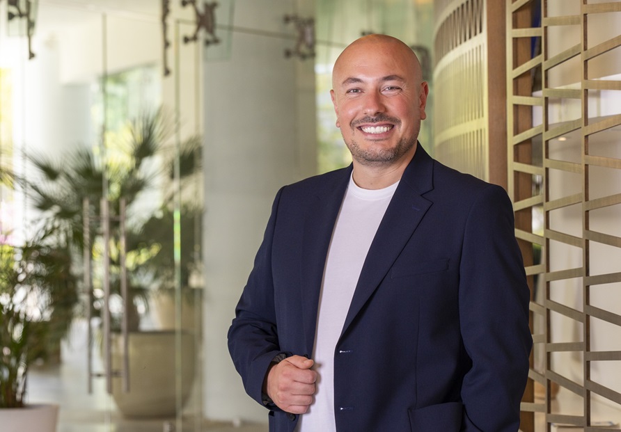 Smiling bald man in a navy blazer and white t-shirt stands in a bright, modern office with glass walls and plants in the background