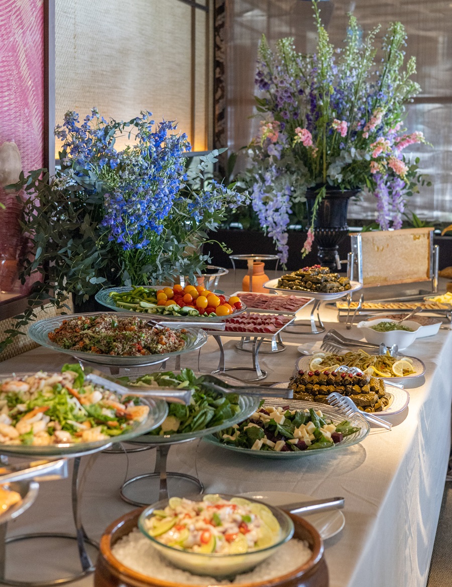 Buffet table with assorted salads and appetizers on glass platters, colorful flowers in blue and purple arrangements in the background.
