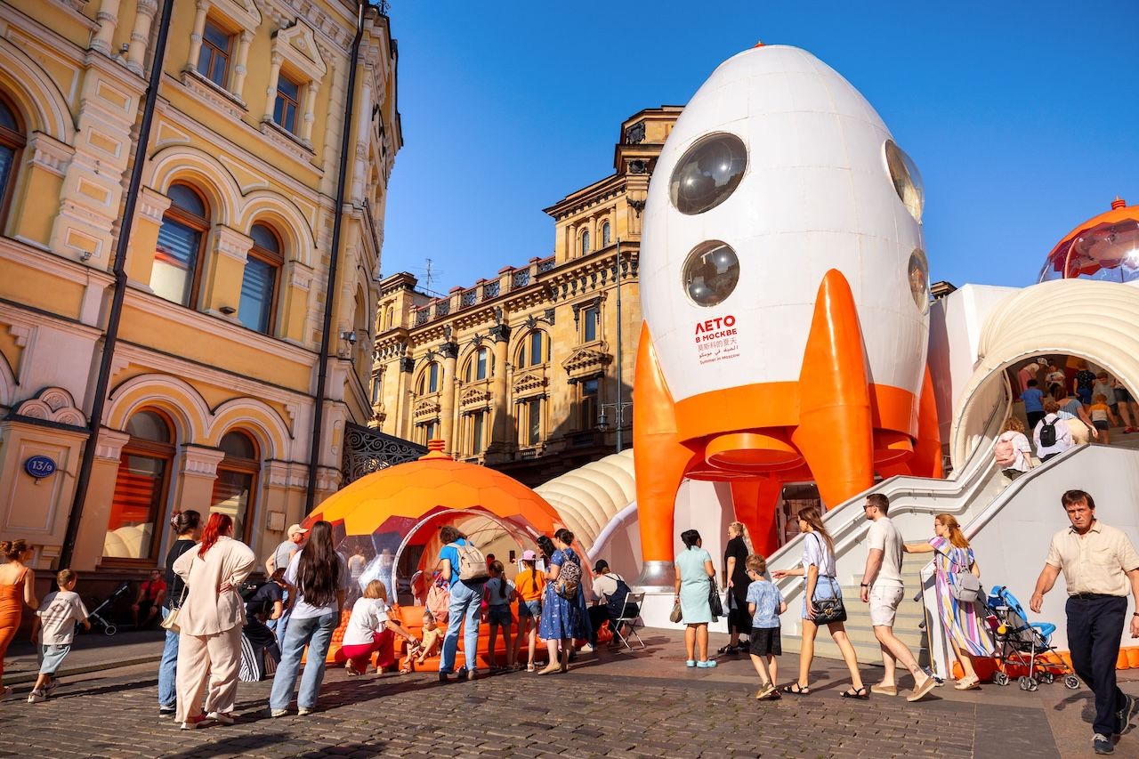 Space-age rocket sculpture with orange boosters on a city street, surrounded by visitors and a curved white entrance, under a bright blue sky.