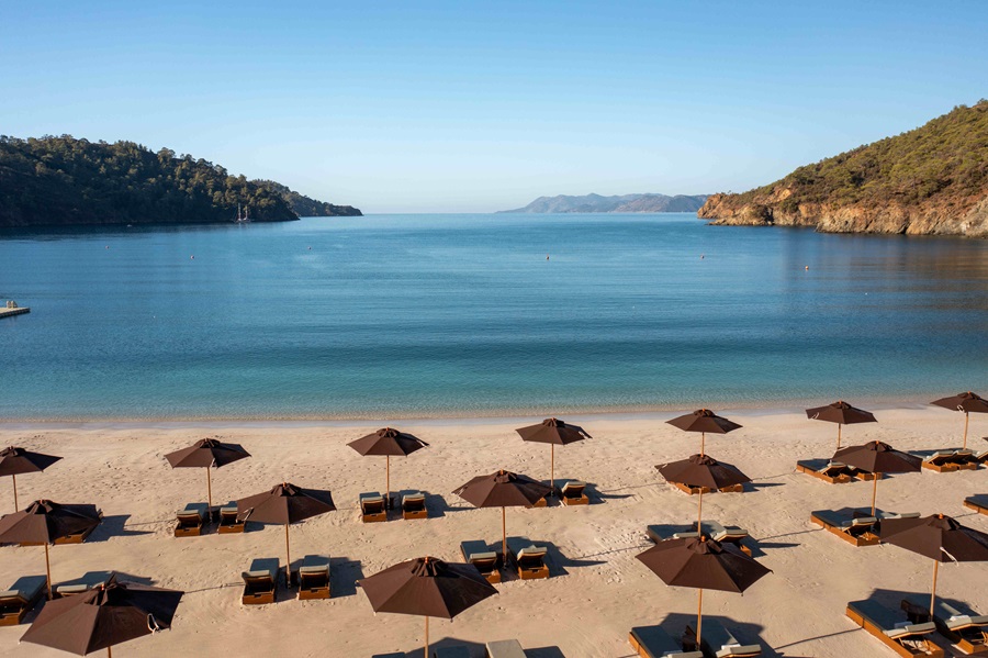 Beach with rows of brown umbrellas and sun loungers on pale sand, calm blue-green water and hills in the distance. - concise, informative alt text for a beach scene.