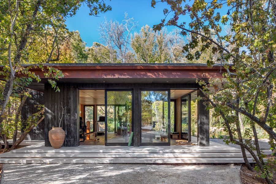 Contemporary single-story home with floor-to-ceiling glass walls, a wooden deck, and surrounding trees; interior gym equipment visible through the left doorway.