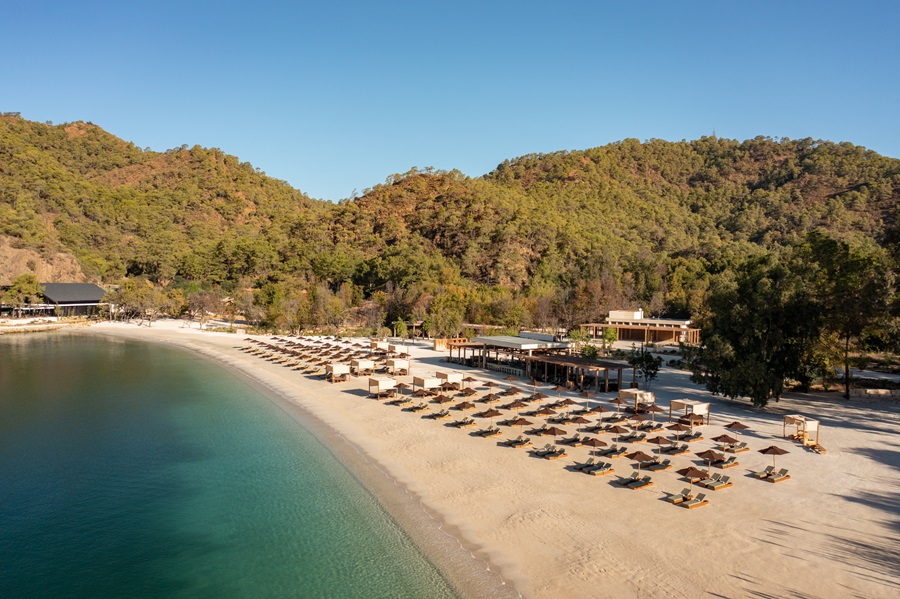 Sandy beach with rows of sun loungers and umbrellas along turquoise water, backed by forested hills and a few buildings.