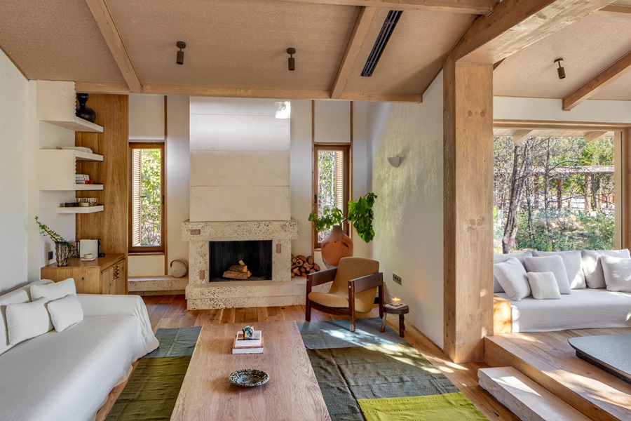 Bright living room with white sofas, wooden beams, and a stone fireplace, plus a sunlit seating area by large windows.