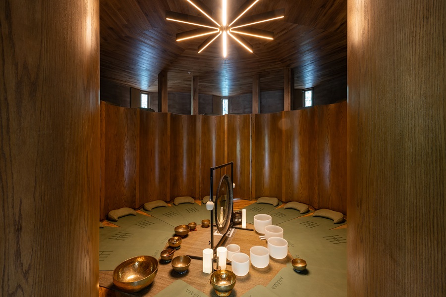 Circular meditation room with wooden curved walls, cushions around a central table holding a gong, singing bowls, and bowls for a sound healing session.