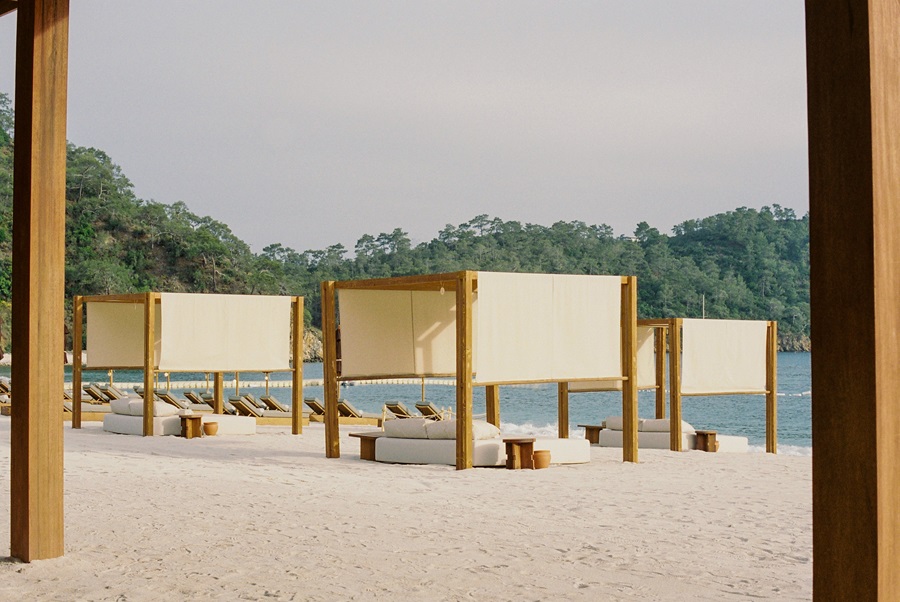 Beach scene with wooden cabanas and white fabric panels, lounge chairs on white sand by calm water.