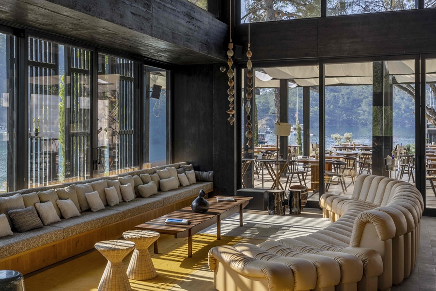 Lounge area with a long beige tufted sofa, low wooden table, and woven stools beside floor-to-ceiling windows overlooking a lakeside patio.
