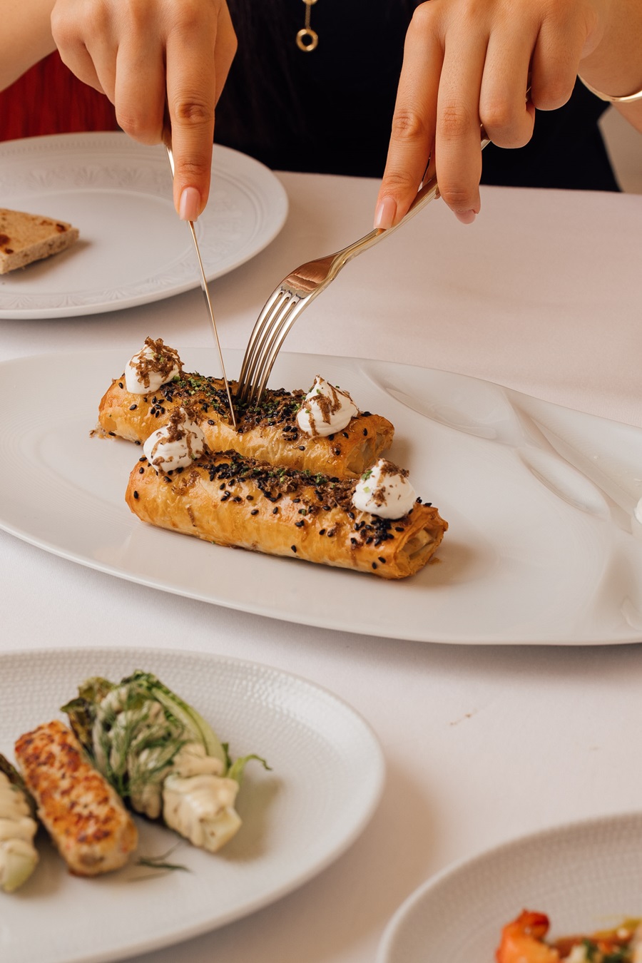 Hands cut two herb-topped puff pastries with dollops of cream on a white plate.
