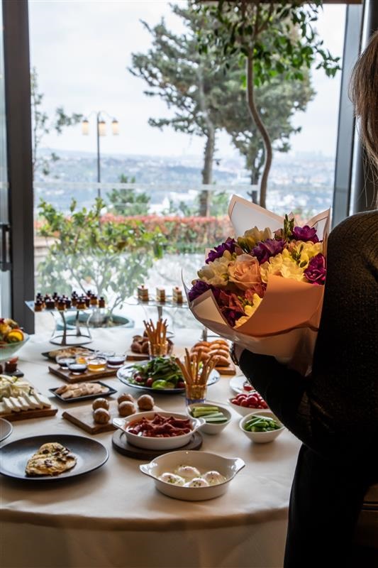 Person holding a bouquet of colorful flowers over a table filled with finger foods at a buffet overlooking a window view.