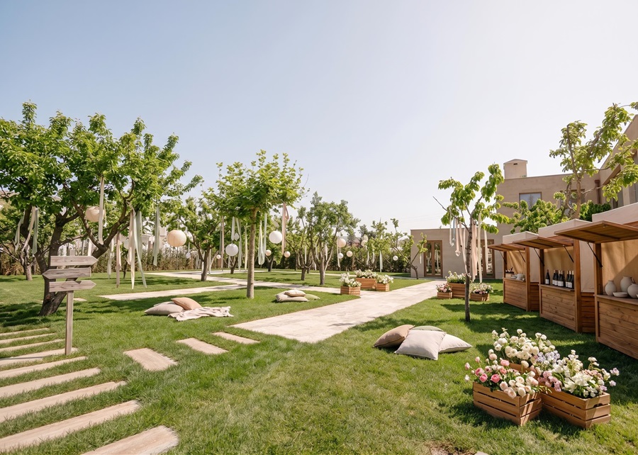 Outdoor garden event setup with trees, hanging balloons, and wooden vendor stalls on a sunny lawn.
