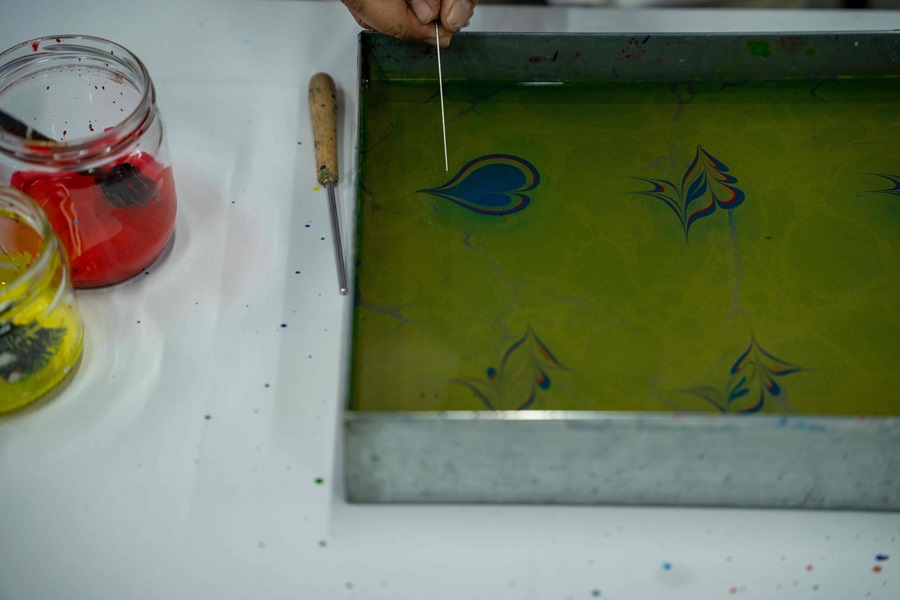 Hand holding a thin metal stylus over a green dye bath in a metal tray; jars of red and yellow paint on the table.