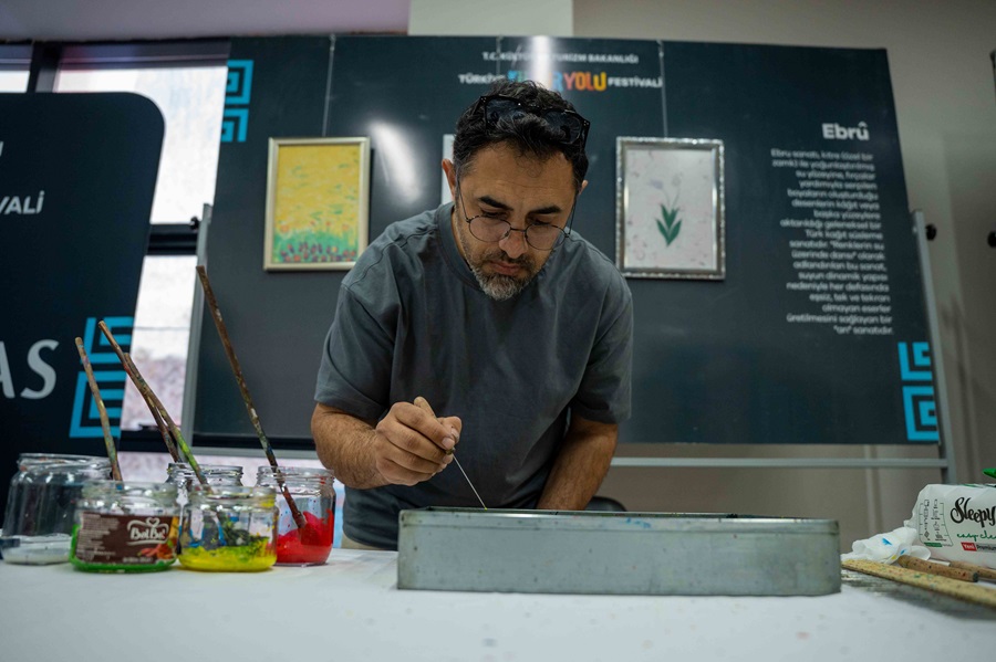 A man with glasses and a gray shirt leans over a table, carefully applying paint with a thin brush amid jars of colorful paints and painting tools in a studio setting.