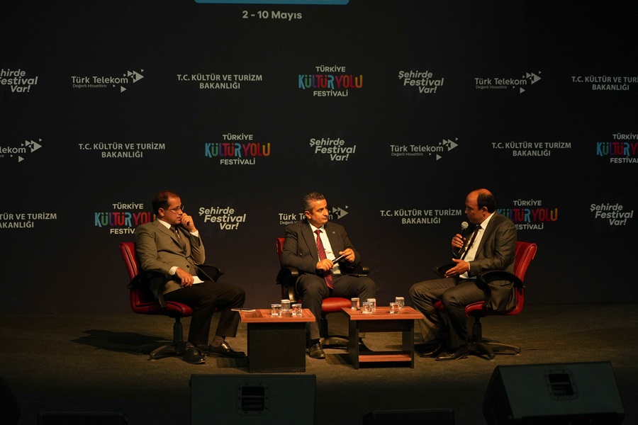 Three men in suits seated on stage in a panel discussion, with a dark backdrop and festival logos behind them.