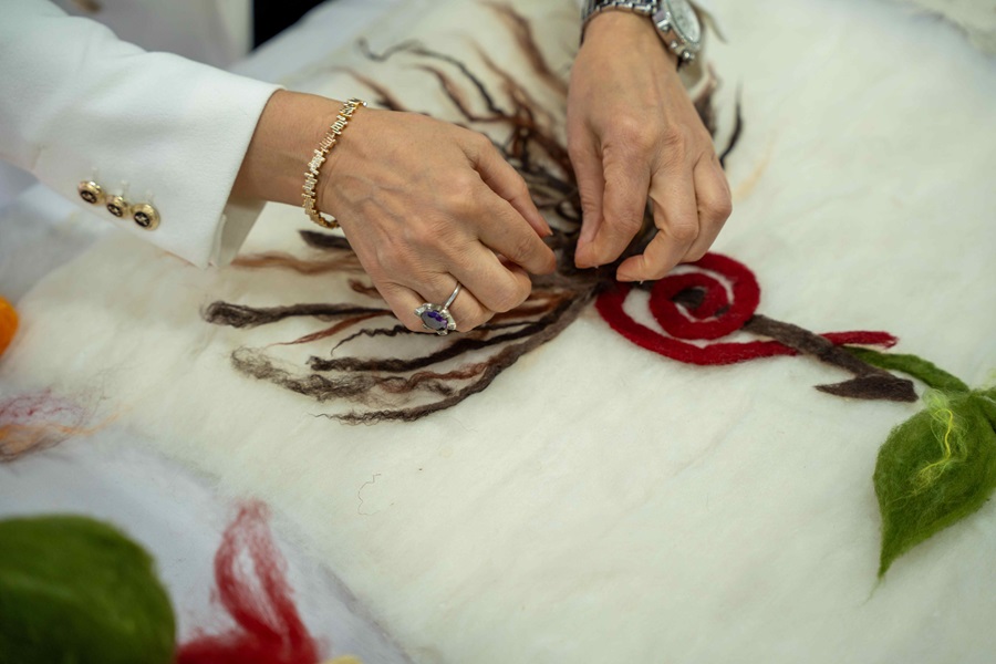 Close-up of two hands needle-felting a red wool rose on white felt with brown stem details.