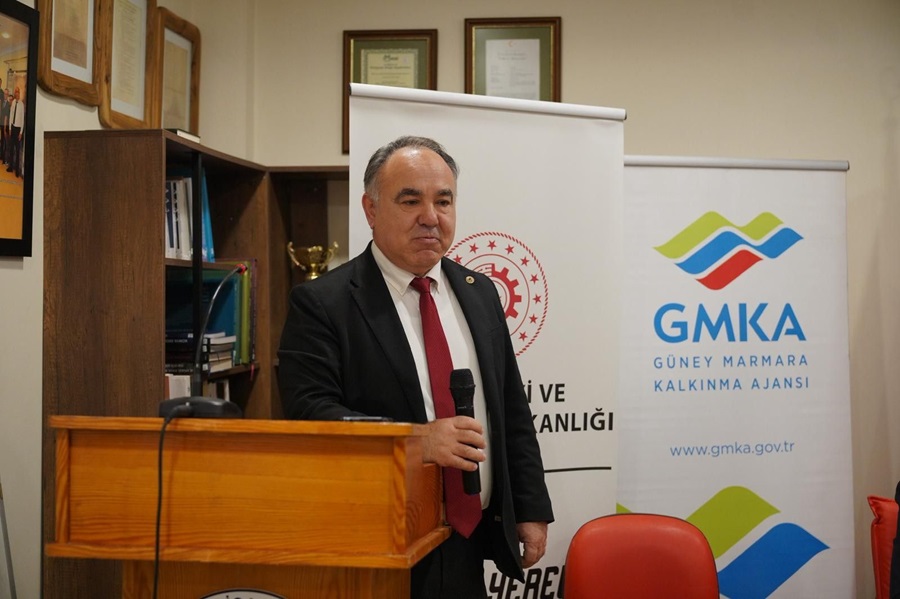 Man in a dark suit and red tie speaks into a handheld microphone at a wooden podium, with banners in the background (GMKA and ministry logo).