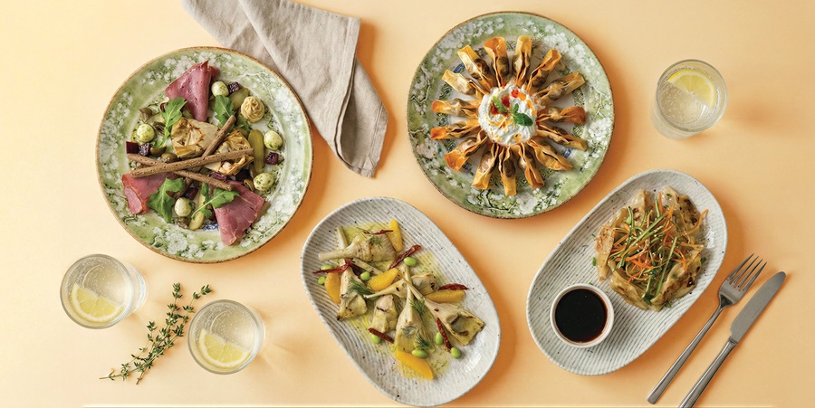 Top-down view of a colorful meal spread: several plated dishes, greens, roasted vegetables, and lemon water on a peach background.