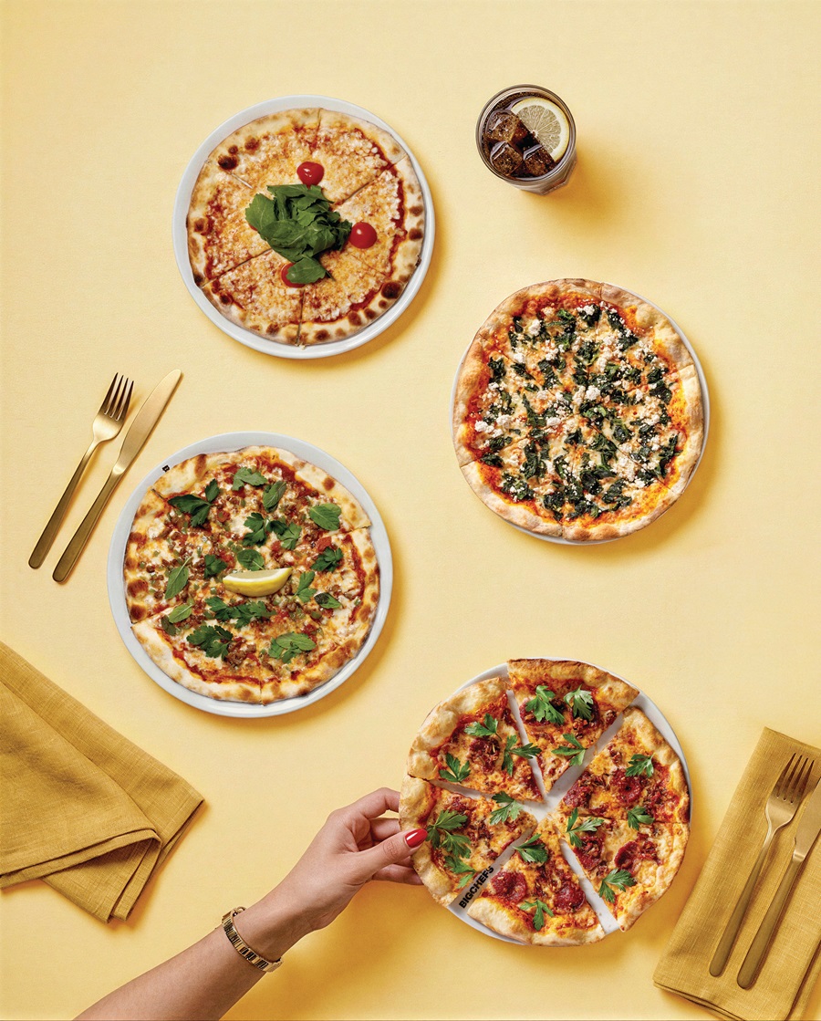 Several pizzas on a yellow table with gold flatware; a hand holds a slice from one pizza in the foreground, others topped with greens and cheese, plus a drink nearby.