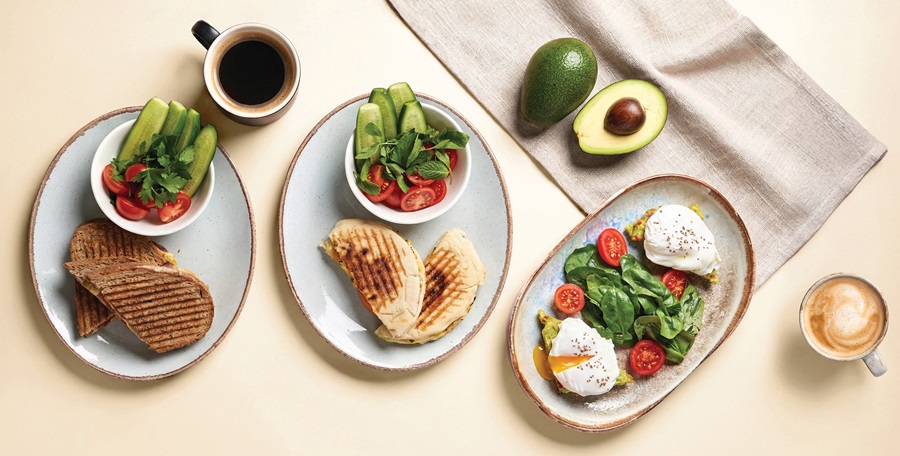 Overhead view of a brunch spread: two grilled sandwiches, a fresh tomato-cucumber salad, avocado halves, and two cups of coffee.