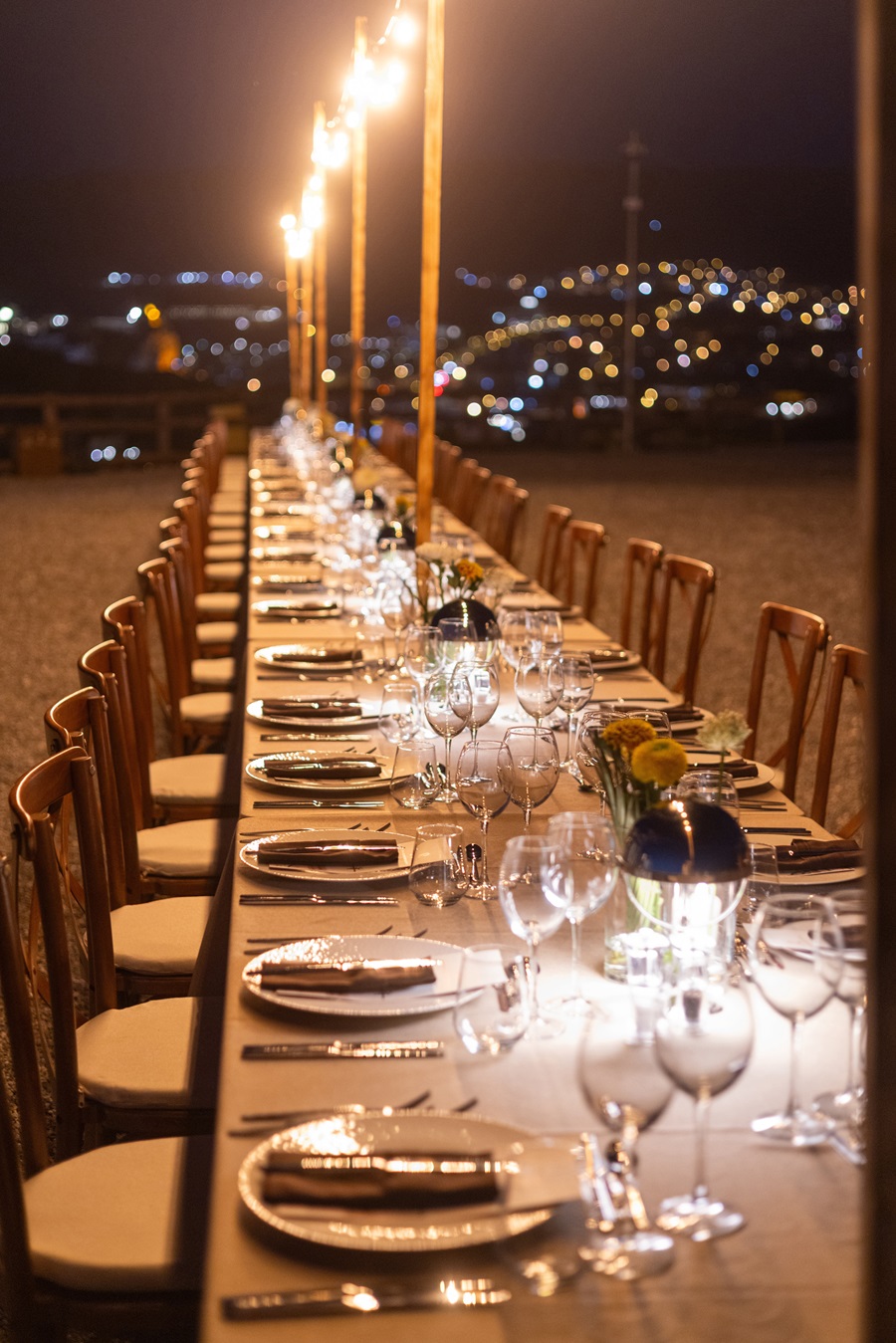 Long outdoor banquet table at night with glassware, plates, and floral centerpieces, city lights in the background.