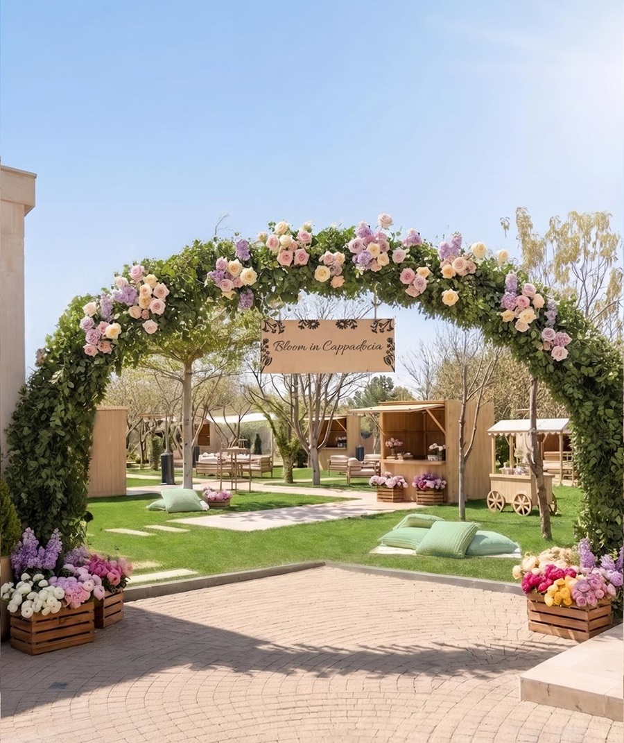 Floral archway with pink and cream roses and greenery over a brick path at a sunny outdoor event, sign reads 'Bloom in Cappadocia'.
