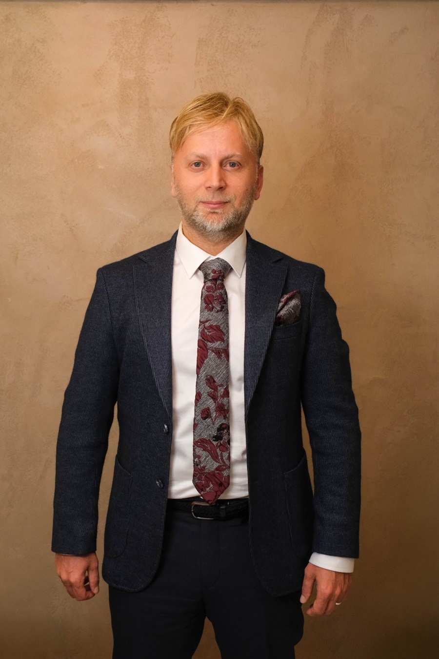 Man in a navy blazer, white shirt, and red-gray patterned tie standing against a beige textured wall.