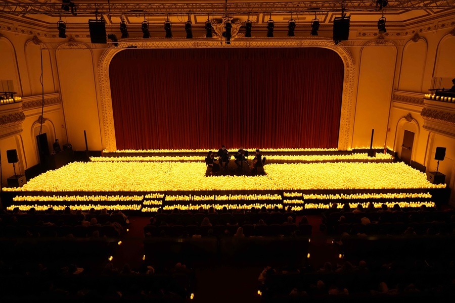 Candles line the stage and seating of a concert hall as a small ensemble gathers for a candlelit performance, with a red curtain backdrop and an attentive audience in dim light.