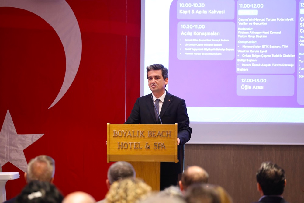 Man in a dark suit delivering a speech at a wooden podium in a hotel conference room, with a Turkish flag backdrop and a projected schedule on screen behind him.
